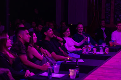 Front-row audience at a small comedy club watching a live show under purple stage lights, seated at candlelit tables with drinks and canned beer.