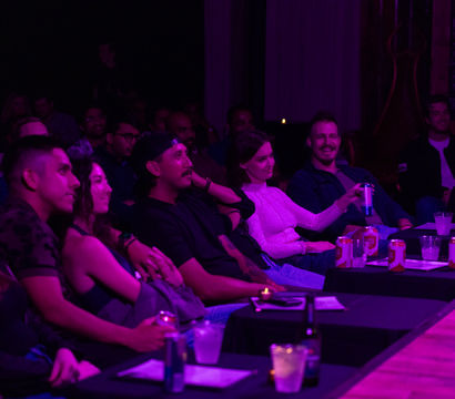 Front-row audience at a small comedy club watching a live show under purple stage lights, seated at candlelit tables with drinks and canned beer.