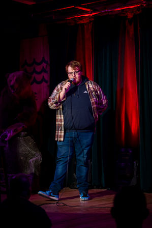 Stand-up comedian on a small comedy club stage holding a microphone, wearing a plaid shirt over a hoodie and sneakers, bathed in red stage lights with curtains, a bear prop to the left and audience silhouettes in the foreground.