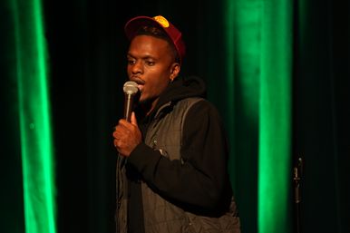 Comedian on stage holding a microphone, wearing a red cap and quilted vest against green vertical stage lights at a live comedy show.