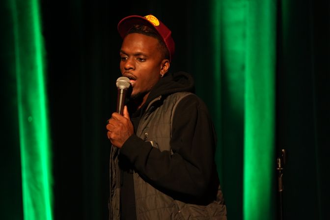 Comedian on stage holding a microphone, wearing a red cap and quilted vest against green vertical stage lights at a live comedy show.