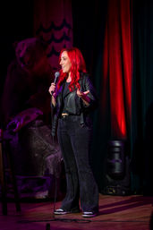 Red-haired stand-up comedian in a leather jacket holding a microphone on a comedy club stage with colorful red and purple lighting.