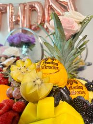 Celebration fruit platter: star-cut golden kiwi, cubed mango, grapes, raspberries, blackberries, pineapple crown and oranges with printed designs, styled with pink roses and rose‑gold party balloons.