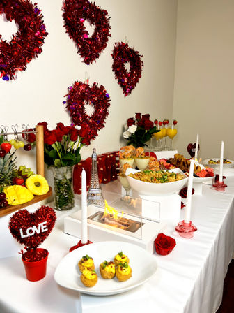 Romantic indoor Valentine’s buffet spread with red tinsel heart wreaths on the wall, red roses, white taper candles, fruit platter, shrimp cocktails, savory appetizers and orange mimosas on a white tablecloth.