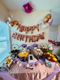 Indoor birthday buffet table at a home party with rose-gold "HAPPY BIRTHDAY" balloons, metallic and confetti balloons, chafing dishes with waffles and casseroles, fresh fruit platter, purple roses and pink table linens.