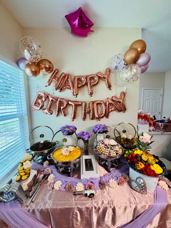 Indoor birthday buffet table at a home party with rose-gold "HAPPY BIRTHDAY" balloons, metallic and confetti balloons, chafing dishes with waffles and casseroles, fresh fruit platter, purple roses and pink table linens.