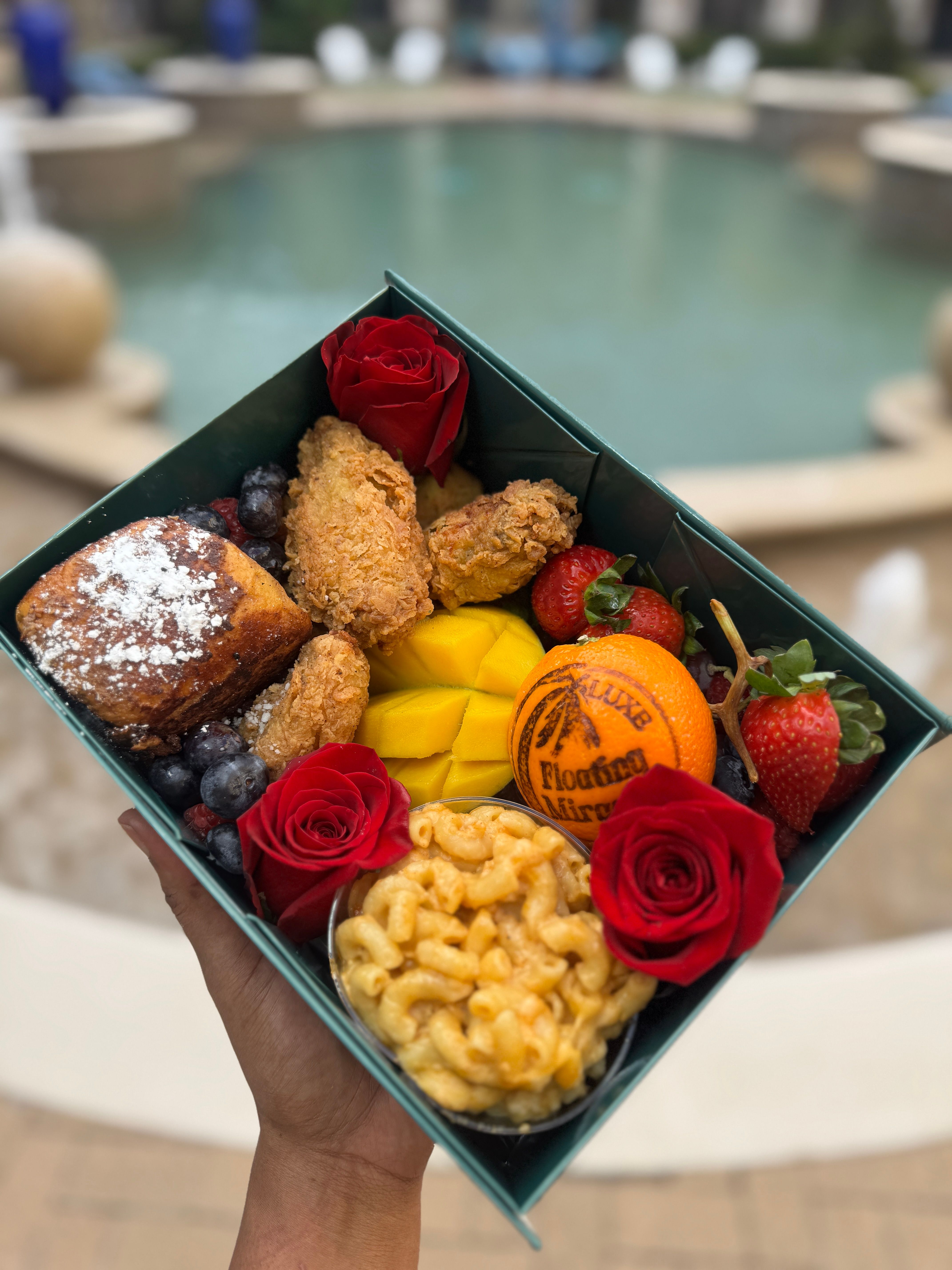 Hand-held poolside brunch box with crispy fried chicken, macaroni and cheese cup, powdered French toast, sliced mango, strawberries, blueberries, a whole orange and red roses.