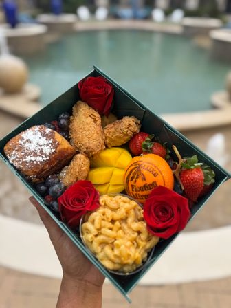 Hand-held poolside brunch box with crispy fried chicken, macaroni and cheese cup, powdered French toast, sliced mango, strawberries, blueberries, a whole orange and red roses.