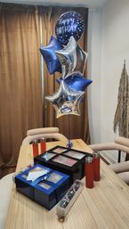 Wooden dining table in a home dining area with a bouquet of blue and silver star-shaped balloons and a round "Happy Birthday" balloon, surrounded by blue and black gift boxes, chocolate-covered strawberries, and red drink tumblers — festive birthday setup.