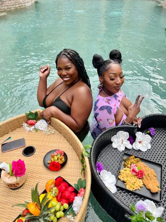 Two women relaxing in a turquoise outdoor pool beside woven floating trays of colorful fruit, flowers, and plated brunch dishes — playful poolside floating breakfast.