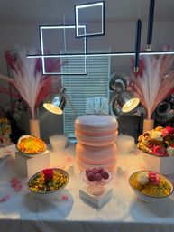 Playful indoor dessert tablescape with a large pink macaron-shaped tiered cake centerpiece, glass pedestal of purple macarons, bowls of colorful snacks topped with roses, pampas grass, LED geometric pendant lights and spotlights over dry-ice fog.