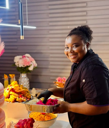 Smiling chef arranging bright pink desserts in a foil tray at an indoor event catering buffet with floral centerpieces and platters of fried and rice dishes