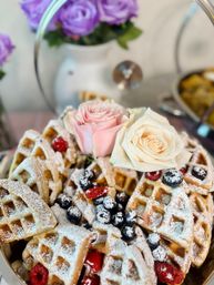 Close-up of golden Belgian waffle quarters dusted with powdered sugar, topped with blueberries and raspberries and garnished with pale pink and cream roses on a tiered serving stand — cheerful brunch dessert.