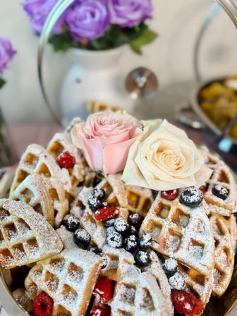 Close-up of golden Belgian waffle quarters dusted with powdered sugar, topped with blueberries and raspberries and garnished with pale pink and cream roses on a tiered serving stand — cheerful brunch dessert.
