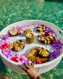 Round white wicker floating brunch tray in a turquoise pool, decorated with pink and purple tropical flowers and a pineapple cup, holding fried chicken, mac and cheese, corn, pastries, grapes and a wine glass while a hand steadies the tray.