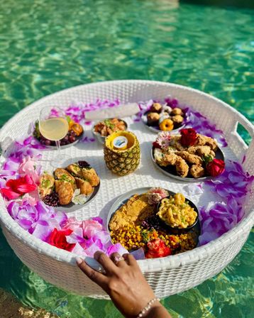 Round white wicker floating brunch tray in a turquoise pool, decorated with pink and purple tropical flowers and a pineapple cup, holding fried chicken, mac and cheese, corn, pastries, grapes and a wine glass while a hand steadies the tray.