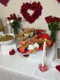 Valentine’s Day brunch table with red heart tinsel and rose bouquets, platter of fried chicken and pink waffles with berries and whipped cream, shrimp cocktail glasses, salad bowl and a lit white taper candle in a pink holder — romantic party spread