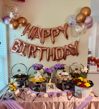 Indoor birthday brunch buffet at home with rose-gold "HAPPY BIRTHDAY" balloons, purple floral accents, chafing dishes of quiche and waffles, fresh fruit platter and champagne flutes.