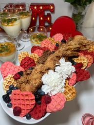 Valentine's brunch platter of crispy fried chicken and heart-shaped pink, red and golden mini waffles garnished with blueberries, raspberries, blackberries and white flowers, with creamy parfaits and a lit red heart marquee in the background.