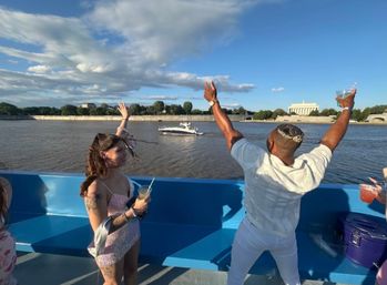 Group of friends on a sunny party boat on the Potomac River in Washington, D.C., holding drinks and cheering toward the Lincoln Memorial across the water under a blue sky.