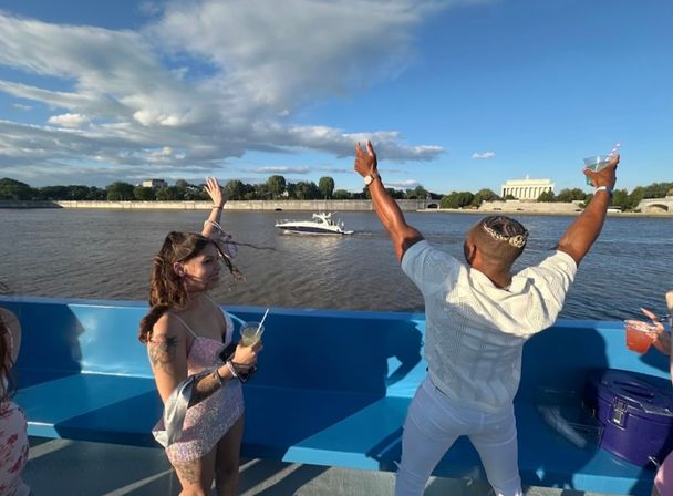 Group of friends on a sunny party boat on the Potomac River in Washington, D.C., holding drinks and cheering toward the Lincoln Memorial across the water under a blue sky.