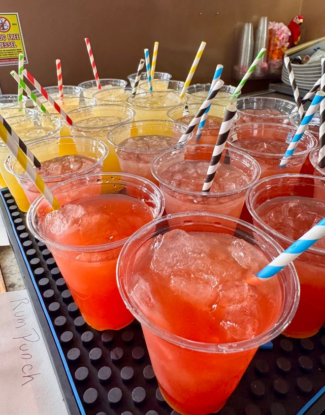 Rows of colorful iced fruit punches in clear plastic cups with striped paper straws on a drink counter — red, orange and yellow flavors ready to serve.