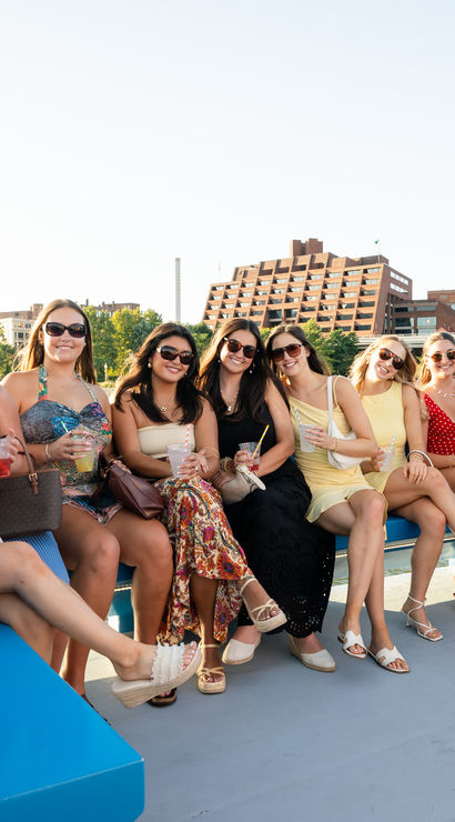Cheerful group of friends on a sunny summer boat cruise along an urban river, smiling and holding colorful cocktails with waterfront buildings and a bridge in the background.