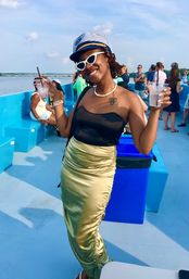 Young woman in a sailor hat and white cat-eye sunglasses holding drinks on a sunny blue boat deck during a summer cruise with water and passengers in the background