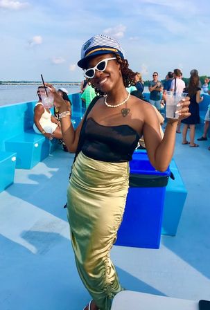 Young woman in a sailor hat and white cat-eye sunglasses holding drinks on a sunny blue boat deck during a summer cruise with water and passengers in the background