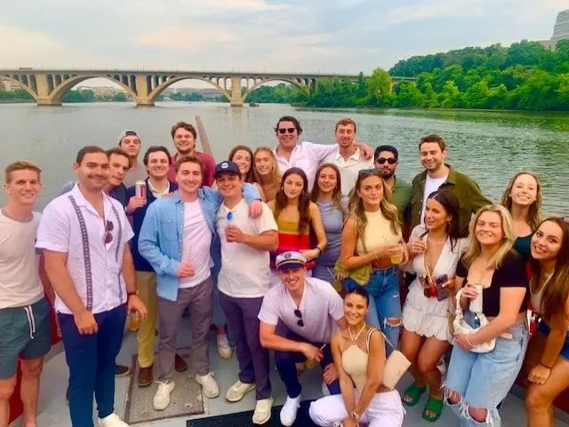 Group of young adults enjoying a summer boat party on a river, posing on the deck with an arched bridge and tree-lined riverbank under a pastel sky.
