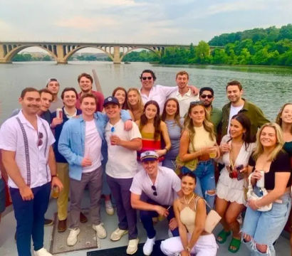 Group of young adults enjoying a summer boat party on a river, posing on the deck with an arched bridge and tree-lined riverbank under a pastel sky.