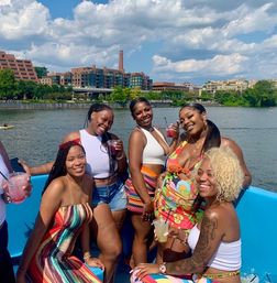 Five friends in colorful summer outfits smiling and holding drinks on a sunny riverboat cruise by an urban waterfront and blue sky.