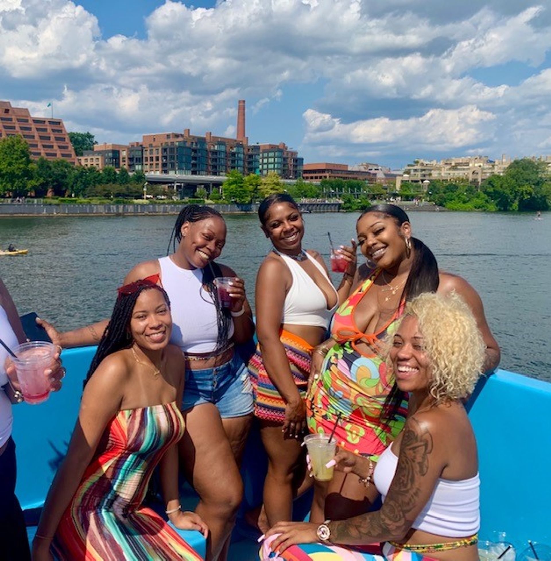 Five friends in colorful summer outfits smiling and holding drinks on a sunny riverboat cruise by an urban waterfront and blue sky.