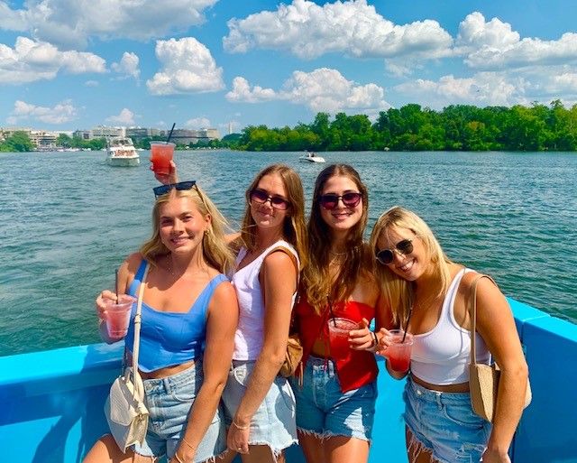 Four friends in sunglasses holding pink cocktails on a bright-blue boat during a sunny summer cruise on a river with green shoreline and city skyline