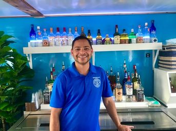 Smiling bartender in a blue polo behind a well-stocked blue-walled bar with rows of liquor bottles on white shelves and a stainless-steel cocktail station