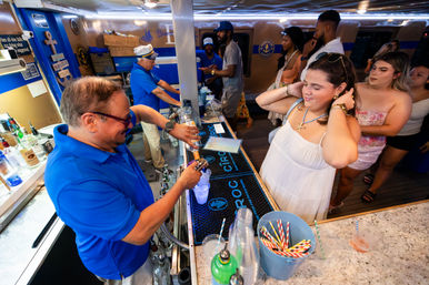 Bartender in a blue polo pouring cocktails at a busy nautical-themed bar on a party boat, smiling patrons in summer outfits waiting at the counter under blue-and-gold decor.