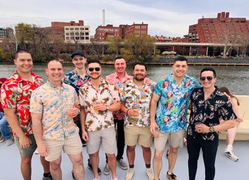 Group of men in colorful Hawaiian shirts smiling on a boat along an urban riverfront with brick buildings and trees in the background.