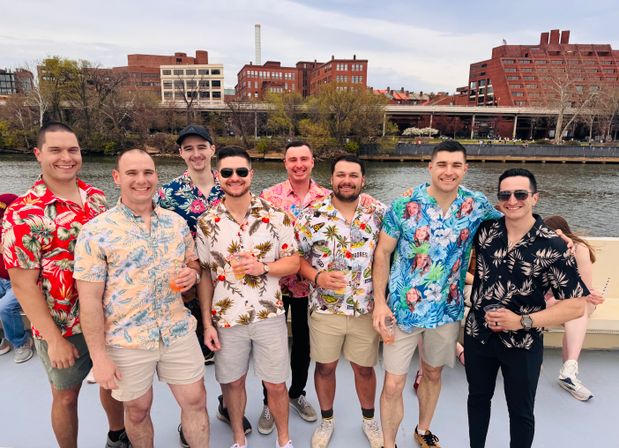 Group of men in colorful Hawaiian shirts smiling on a boat along an urban riverfront with brick buildings and trees in the background.