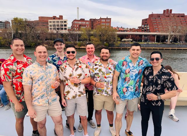 Group of eight men in colorful Hawaiian shirts smiling on a riverboat deck with drinks, urban brick waterfront and trees in the background on a cloudy spring day.