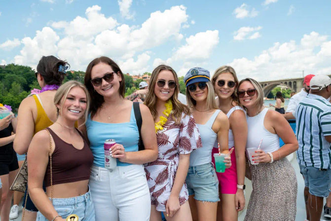 Six smiling women in summer outfits and sunglasses holding colorful drinks on a sunny boat party by a river, with a stone arched bridge and blue sky; one wears a captain's hat and a lei.