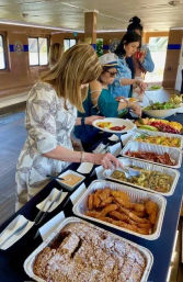 Onboard a scenic cruise boat, guests serve themselves from a buffet table with trays of fried chicken strips, savory quiche, mixed fruit, salads and pastries beside windows with water views.