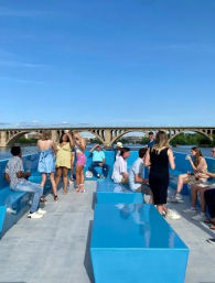 Young adults socializing on a bright blue-deck river cruise boat on a sunny day, with an arched stone bridge and waterfront in the background.