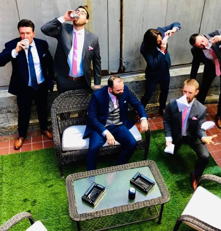 Overhead shot of well-dressed men in suits with pink ties drinking canned beers and tossing paper on a small urban patio with wicker seating and green artificial turf.