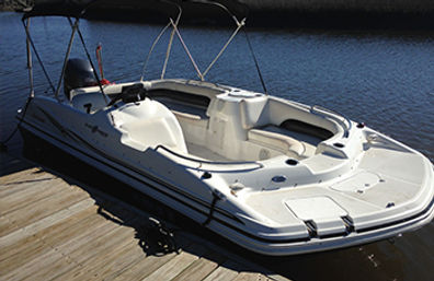 Sunlit white deck boat with bimini top and outboard motor docked at a wooden pier on calm lake water