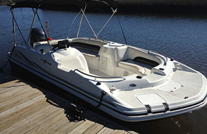 Sunlit white deck boat with bimini top and outboard motor docked at a wooden pier on calm lake water