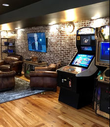 Cozy home basement game room with exposed brick wall, wall-mounted TV showing a news program, brown leather armchairs on a rug, hardwood floor and retro arcade cabinets.