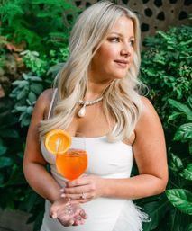 Elegant blonde woman in a white dress holding an orange cocktail with an orange slice, wearing a pearl pendant necklace, posing against lush green garden foliage — outdoor summer portrait.