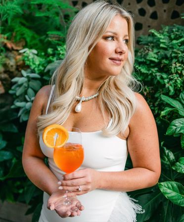 Elegant blonde woman in a white dress holding an orange cocktail with an orange slice, wearing a pearl pendant necklace, posing against lush green garden foliage — outdoor summer portrait.