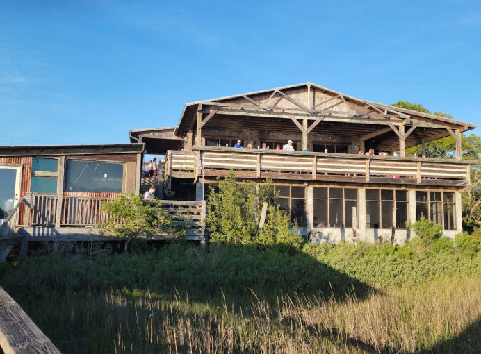 Sunlit rustic two-story coastal wooden building with an open upper deck of diners, screened lower porch and marsh grasses in the foreground under a clear blue sky.