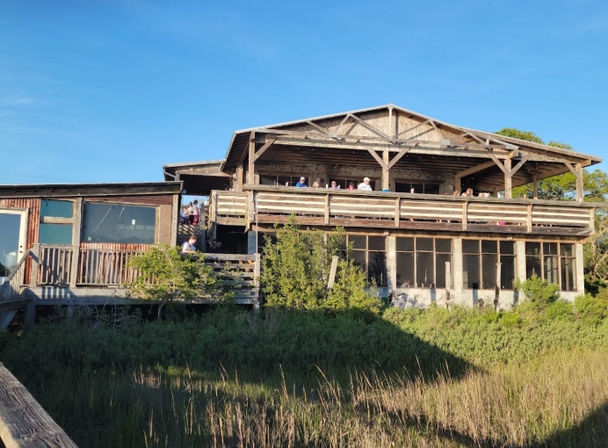 Sunlit rustic two-story coastal wooden building with an open upper deck of diners, screened lower porch and marsh grasses in the foreground under a clear blue sky.